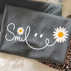 Puede incluir: Una sudadera gris con la palabra "Smile" en escritura blanca, con acentos de margaritas. La "i" de smile se reemplaza con una margarita. Una segunda margarita está en el lado derecho de la palabra. La sudadera está doblada.