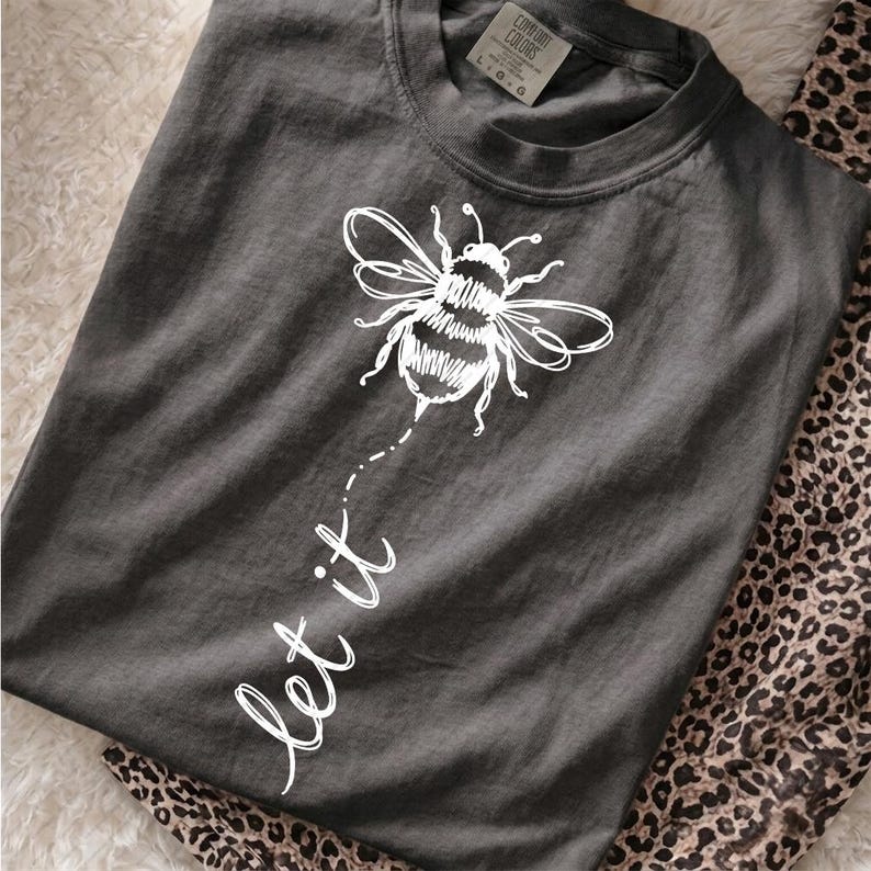 Op de afbeelding: Een antracietgrijs T-shirt met ronde hals voorzien van een witte lijntekening van een hommel boven de handgeschreven tekst "let it" die met een stippellijn vanaf de bij naar beneden loopt op de voorzijde van het kledingstuk.