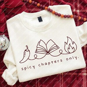 May include: White crewneck sweatshirt with a graphic design in dark red. The design features a chili pepper, an open book, and flames, connected by a line, with the text "spicy chapters only."