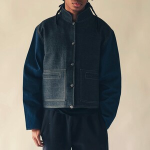 May include: A denim jacket with a stand-up collar and button closure. The jacket features a two-tone design, with darker denim on the body and contrasting blue sleeves. Two square pockets are visible on the front. The jacket is a modern take on a classic style.