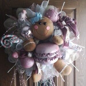 以下が含まれることがあります： 茶色のドアに、ピンクと白のマカロン、キャンディーケーン、ピンクと白のリボンをつけたジンジャーブレッドマンの人形。白いメッシュとグリッターで飾られています。