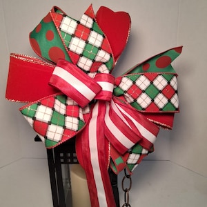 May include: A decorative multi-layered bow featuring a mix of red velvet, green with red polka dots, and a white, green, and black plaid pattern. The bow includes long streamers with red and white stripes and is attached to a black metal lantern.