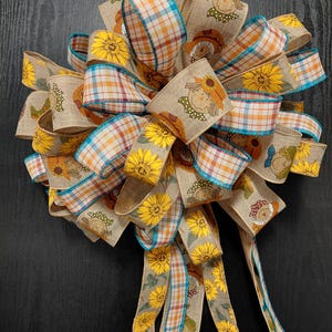 May include: A decorative bow made from multiple ribbons featuring a tan burlap texture. The ribbons are patterned with yellow sunflowers, scarecrow faces, and a multi-colored plaid design with blue edges. The bow has several long, trailing tails.