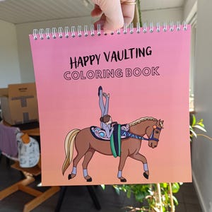 Op de afbeelding: Een roze kleurboek met een spiraalbinding. De omslag toont een cartoonillustratie van een persoon die voltigeert op een bruin paard. De titel van het boek is "Happy Vaulting Coloring Book".
