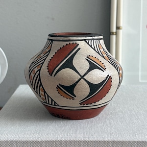May include: A small, handcrafted ceramic pot with a rounded body and a narrow neck. The pot features a cream-colored background with geometric designs in black, orange, and brown. The pot is sitting on top of a book with the word "gestalten" printed on it.