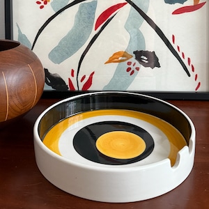 May include: A white ceramic ashtray with a black, yellow, and white circular design. The ashtray has a curved rim and a small notch for resting cigarettes. A wooden bowl and a framed abstract painting are in the background.