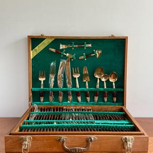 May include: A wooden flatware storage case with a green velvet interior containing a large set of gold-toned utensils with dark brown handles. The set includes forks, knives, and spoons, some wrapped in plastic, with a label reading Anan's.