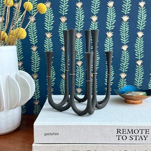 May include: A black metal candelabra with seven candle holders. The candelabra is on top of two books, one with the title "gestalten" and the other with the title "REMOTE TO STAY". A white vase with yellow flowers is to the left.