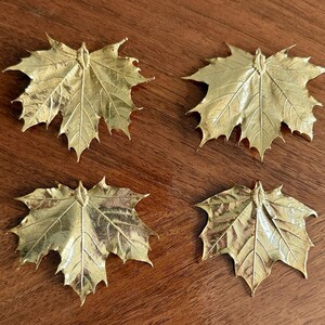 May include: Four gold-colored maple leaf decorations arranged on a wooden surface. Each leaf has detailed veining and a textured surface, creating a realistic appearance. The leaves are likely decorative items.