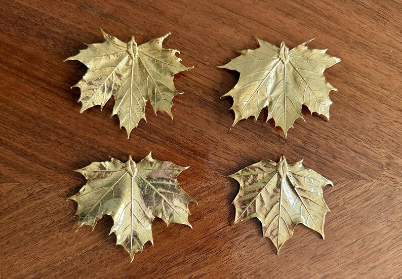 May include: Four gold-colored maple leaf decorations arranged on a wooden surface. Each leaf has detailed veining and a textured surface, creating a realistic appearance. The leaves are likely decorative items.