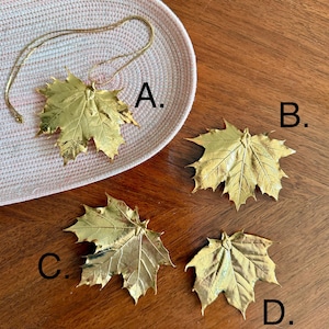 May include: A collection of gold-colored maple leaf pendants and charms. One pendant is on a gold chain, resting on a woven pink and white tray. Three additional leaf charms are displayed on a wooden surface. The leaves have detailed veining.