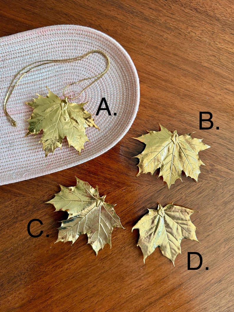 May include: A collection of gold-colored maple leaf pendants and charms. One pendant is on a gold chain, resting on a woven pink and white tray. Three additional leaf charms are displayed on a wooden surface. The leaves have detailed veining.