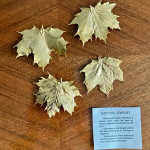May include: Four gold-colored maple leaf ornaments are arranged on a wooden surface. The leaves have intricate vein details and a metallic sheen. A small blue card with the text "NATURAL JEWELRY" is also present.