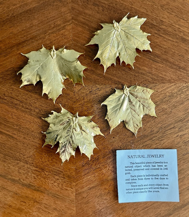 May include: Four gold-colored maple leaf ornaments are arranged on a wooden surface. The leaves have intricate vein details and a metallic sheen. A small blue card with the text "NATURAL JEWELRY" is also present.