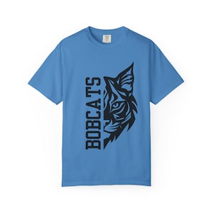 May include: Light blue t-shirt featuring a black graphic of a bobcat's face and the word "BOBCATS" in black vertical text. The shirt is made of a soft material and has short sleeves.
