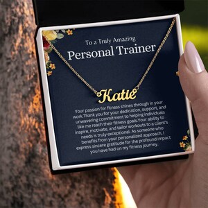 May include: A gold necklace with the name "Katie" on it, inside a blue box with a floral design. The box has a message that reads "To a Truly Amazing Personal Trainer".