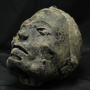 Pode incluir: Uma escultura escura e desgastada de uma cabeça humana com uma superfície preta fosca e texturizada. Os traços faciais incluem olhos profundos, um nariz proeminente e lábios ligeiramente entreabertos. A superfície está coberta de areia.