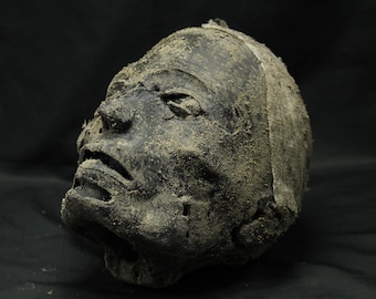 Può includere: Una scultura scura e usurata di una testa umana con una superficie nera opaca e ruvida. I tratti del viso includono occhi infossati, un naso prominente e labbra leggermente socchiuse. La superficie è ricoperta da un fine residuo sabbioso.