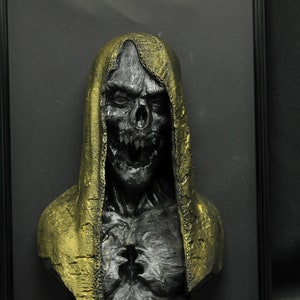 May include: A 3D printed sculpture of a hooded figure with a skull-like face, painted in black and gold. The figure is framed and appears to be a horror or fantasy themed art piece.