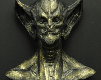 May include: A detailed bust of a creature with a humanoid face, large pointed ears, and a textured surface. The sculpture is primarily dark with gold highlights, set against a black background.