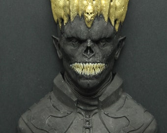May include: A black and gold bust sculpture of a creature with a crown, sharp teeth, and a skull on the crown.