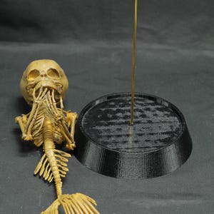 May include: A tan-coloured mermaid skeleton model, featuring a skull, rib cage, and a fan-shaped tail. A black, circular base and a gold rod are also present. The model is likely for Halloween or a nautical theme.