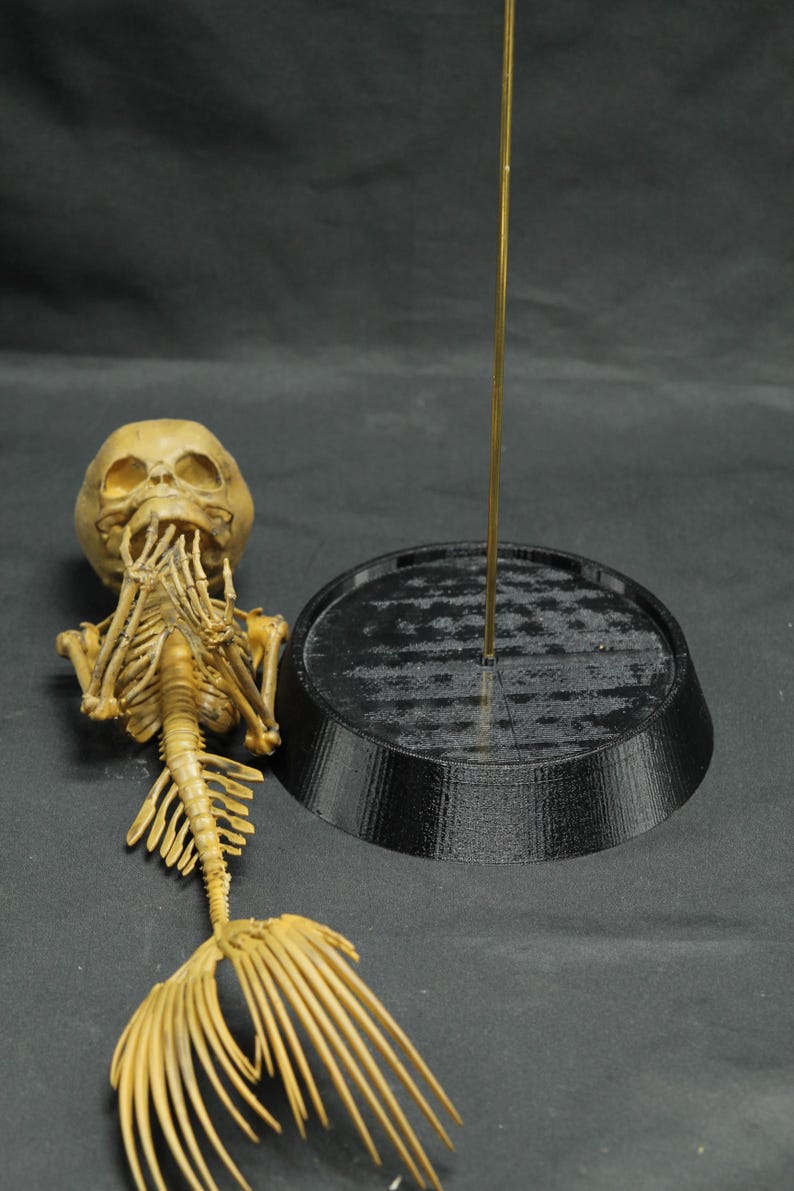 May include: A tan-coloured mermaid skeleton model, featuring a skull, rib cage, and a fan-shaped tail. A black, circular base and a gold rod are also present. The model is likely for Halloween or a nautical theme.
