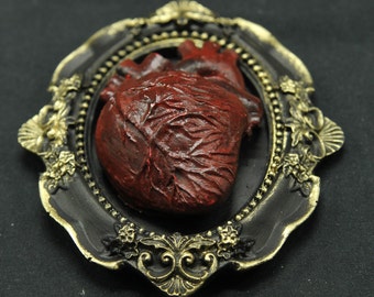 May include: A red anatomical heart in a black and gold ornate frame. The frame has a decorative design with floral motifs.