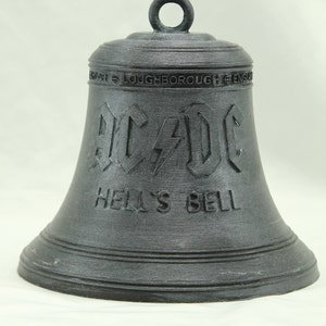 Op de afbeelding: Een zwarte metalen bel met de woorden "AC/DC" en "Hell's Bell" erop gegraveerd. De bel heeft een lus aan de bovenkant om op te hangen.