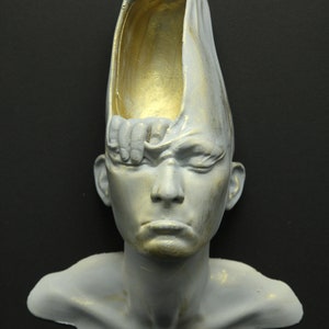 May include: A white plaster sculpture of a person's head with a hand reaching out from the top of the head. The hand is holding a piece of fabric that is draped over the head. The head is painted with gold accents.