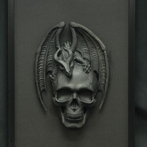 May include: A black framed wall decor featuring a 3D sculpture of a black dragon perched on top of a black skull.