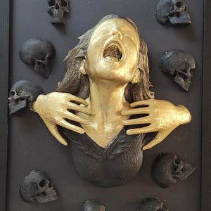 Puede incluir: Una escultura dorada de una mujer con las manos alrededor del cuello, gritando. La escultura está rodeada de cráneos negros.