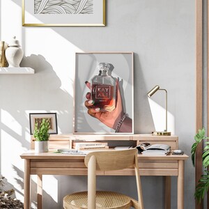 May include: A framed print featuring a perfume bottle held in a hand with a silver bracelet. The bottle is labeled "KAY ALI" and has a clear, faceted top. The artwork is displayed on a wooden desk with a gold lamp and a small potted plant.