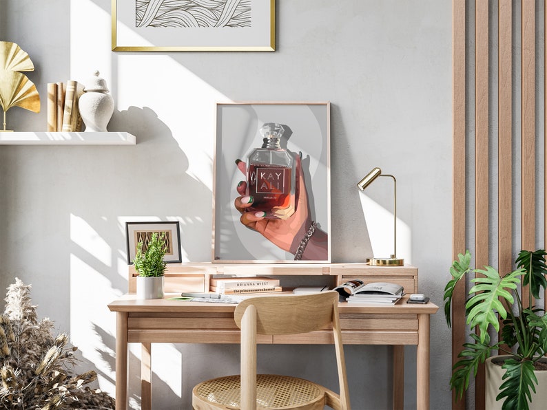 May include: A framed print featuring a perfume bottle held in a hand with a silver bracelet. The bottle is labeled "KAY ALI" and has a clear, faceted top. The artwork is displayed on a wooden desk with a gold lamp and a small potted plant.