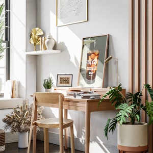 Pu&ograve; includere: Un ambiente di lavoro domestico con una scrivania in legno, una sedia e una stampa incorniciata con il testo "my cup runneth over". La scrivania ha una lampada, una pianta in vaso e un laptop. La stanza ha una grande finestra, una pianta in vaso e uno scaffale con oggetti decorativi.