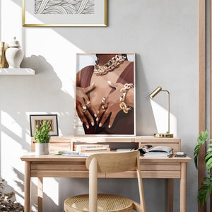 May include: A framed print of a person wearing gold jewelry, including a necklace, bracelet, and rings. The artwork is displayed on a wooden desk with a gold lamp, a small plant, and a wooden chair. A white shelf holds decorative items.