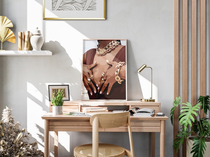 May include: A framed print of a person wearing gold jewelry, including a necklace, bracelet, and rings. The artwork is displayed on a wooden desk with a gold lamp, a small plant, and a wooden chair. A white shelf holds decorative items.