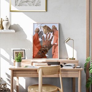 May include: An art print featuring a hand adorned with gold jewelry and rings, set against a backdrop of orange and blue. The print is framed and displayed on a wooden desk with a gold lamp and a small plant.
