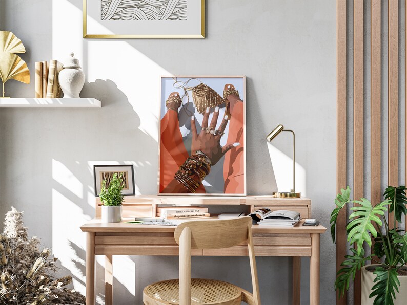 May include: An art print featuring a hand adorned with gold jewelry and rings, set against a backdrop of orange and blue. The print is framed and displayed on a wooden desk with a gold lamp and a small plant.