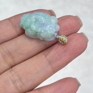May include: A carved jade pendant in the shape of a mythical creature, featuring translucent light green and lavender hues. The pendant is attached to a small, decorative bail made of silver-colored cord wrapped with thin gold-toned wire.