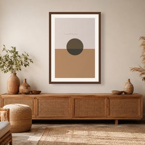 May include: A framed wall art print featuring a split-color background with a light beige upper half and a tan lower half. A dark charcoal circle is centered at the horizon line. The print is enclosed in a dark brown frame with a white mat border.