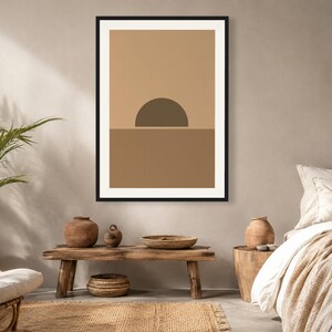May include: A framed wall art print featuring a minimalist graphic design of a dark brown semi-circle sun rising over a solid medium brown horizon line, set against a light tan background, enclosed within a thin black rectangular frame.