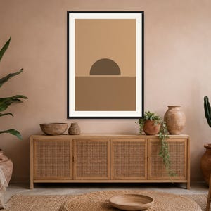 May include: A framed wall art print featuring a minimalist graphic design of a dark brown semi-circle sun positioned above a solid medium brown horizontal line, set against a light tan background within a thin black rectangular frame.