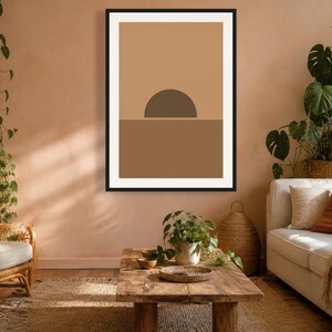 May include: A framed wall art print featuring a minimalist graphic design of a dark brown semi-circle sun rising over a solid medium brown horizon line, set against a light tan background, enclosed within a thin black rectangular frame.