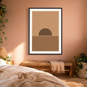 May include: A framed wall art print featuring a minimalist graphic design of a dark brown semi-circle sun rising over a solid medium brown horizon line, set against a light tan background within a thin black frame on a solid wall.
