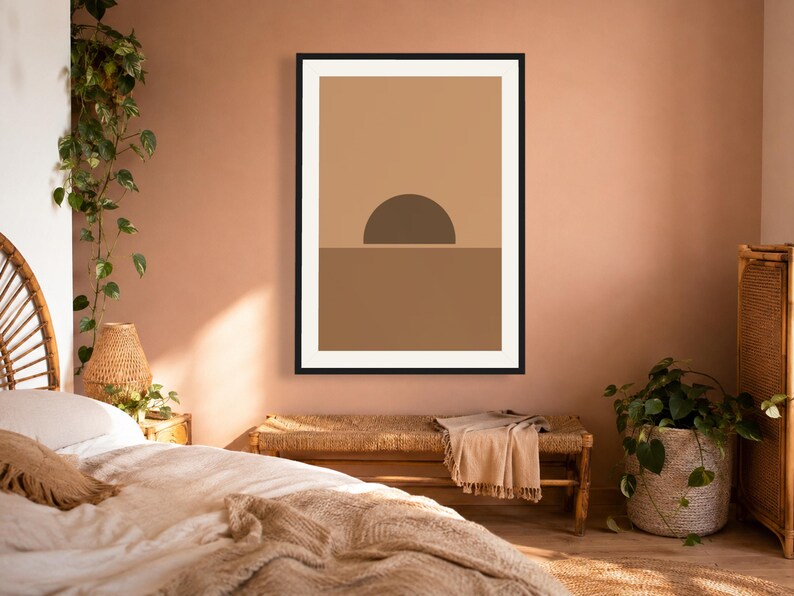 May include: A framed wall art print featuring a minimalist graphic design of a dark brown semi-circle sun rising over a solid medium brown horizon line, set against a light tan background within a thin black frame on a solid wall.