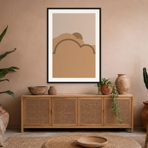 May include: A framed wall art print featuring a minimalist abstract landscape with layered, rounded shapes in shades of tan, beige, and brown. The composition includes a semi-circular sun element partially obscured by cloud-like forms in a flat design.