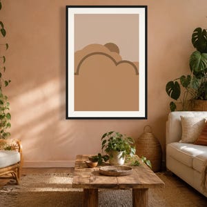 May include: A framed wall art print featuring a graphic illustration of layered abstract shapes in shades of tan, beige, and brown. The design includes curved, cloud-like forms and a semi-circular sun element set against a solid light background.