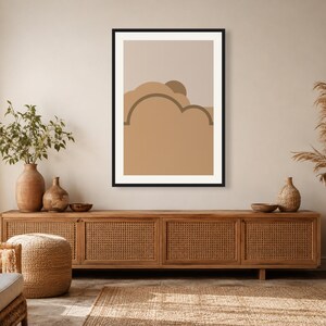 May include: A framed wall art print featuring a minimalist abstract landscape with layered shapes in shades of beige, tan, and brown. The composition includes a semi-circular sun element partially obscured by rounded cloud-like forms in a flat design.