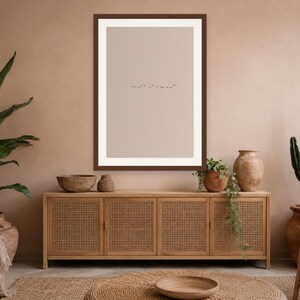 May include: A rectangular wall art print featuring a minimalist beige background with a small, centered line of tiny black birds in flight, enclosed within a dark brown frame, displayed above a wooden sideboard with four cane-paneled doors.